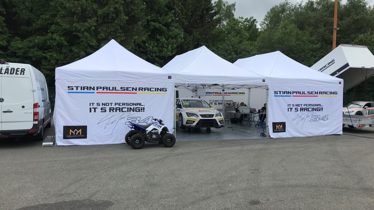 Biały namiot reklamowy od Stian Paulsen Racing, z samochodami wyścigowymi i sprzętem wokół niego. Namiot jest ozdobiony logotypami i tekstami związanymi z wyścigami. Hallgruppen dostarczył ten namiot.