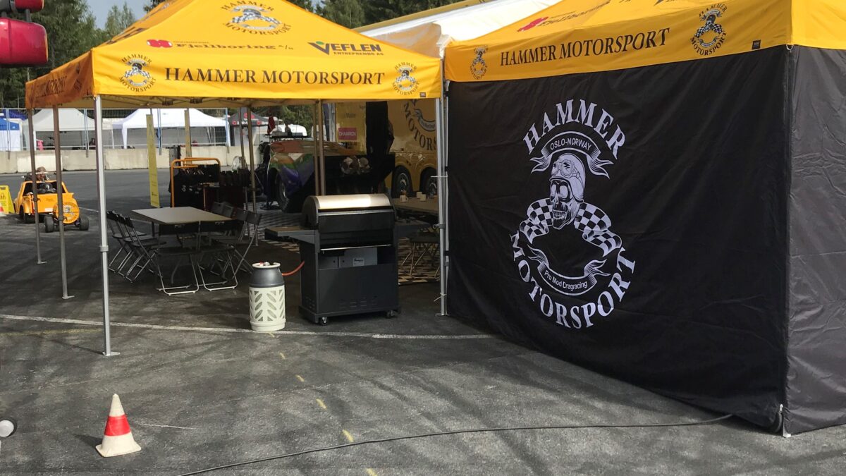 Dwa namioty reklamowe z brandingiem Hammer Motorsport, rozstawione na wydarzeniu sportów motorowych. Namioty są jasne, żółte z czarnymi i czerwonymi detalami.