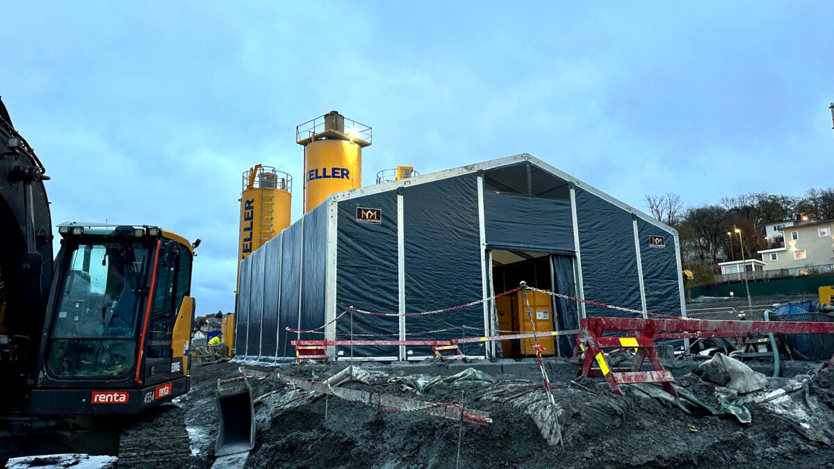 Hala przemysłowa od Hallgruppen ustawiona na placu budowy, z koparką na pierwszym planie i wysokimi żółtymi silosami w tle.