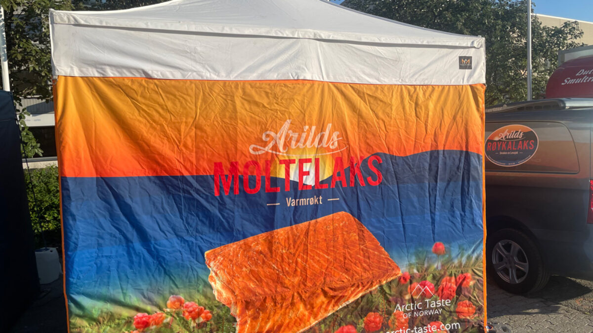 Namiot reklamowy z napisem „Arilds Moltelaks”, wędzonym łososiem i obrazami maliny moroszki. Namiot jest biały z kolorowym tłem i stoi na wydarzeniu, z samochodem w tle. Hallgruppen dostarczył ten namiot.