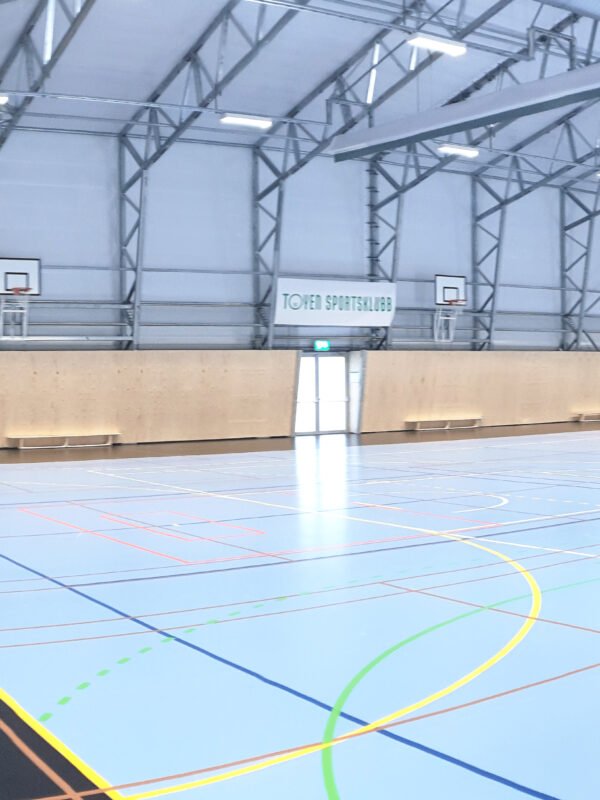 Wnętrze dużej hali sportowej z niebieską podłogą i liniami sportowymi. Hallgruppen zbudował tę halę.